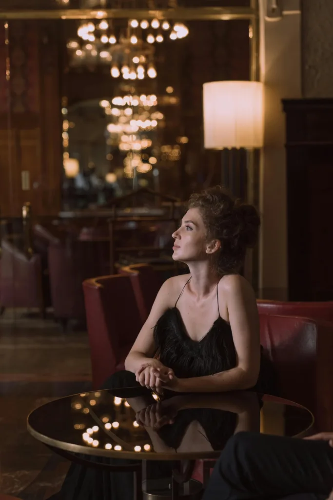 Jovem com um vestido de noite preto sentada a uma mesa no saguão de um luxuoso hotel, olhando para cima, em direção a um grande lustre.