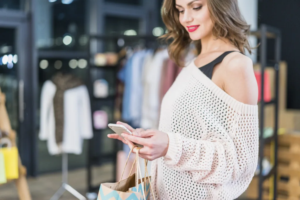 Mulher sorridente segurando sacolas de compras enquanto utiliza seu smartphone em uma loja de roupas.